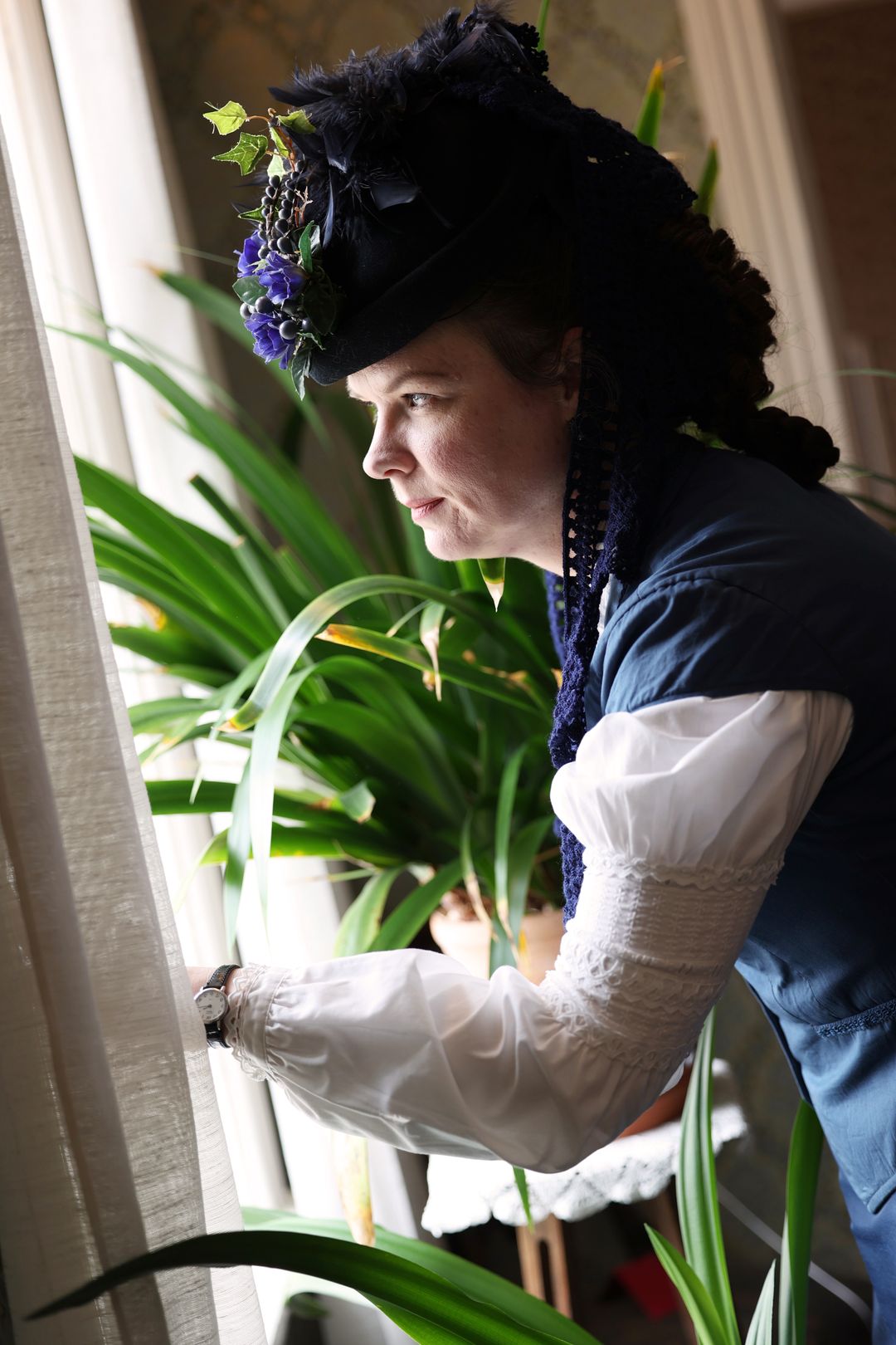 Taina Parikka kurkistaa verhojen välistä ulos ikkunasta 1880-luvun asussaan. Kuva on otettu Parikasta katsoen sivusuunnassa ja on puolikuva. Taustalla huonekasvi.
