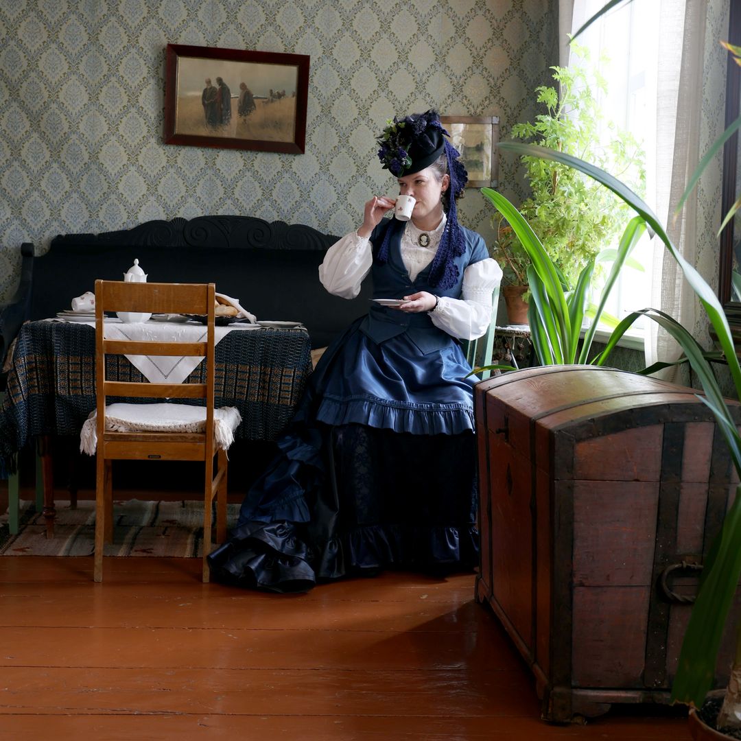 Museonhoitaja Taina Parikka istuu Kauppiaantalomuseon salissa, jossa on huonekasveja, tauluja ja kahvipöytä tuoleineen ja sohvineen. Parikka istuu pöydän äärellä ja juo teetä vanhanaikaisesta teekupista.
