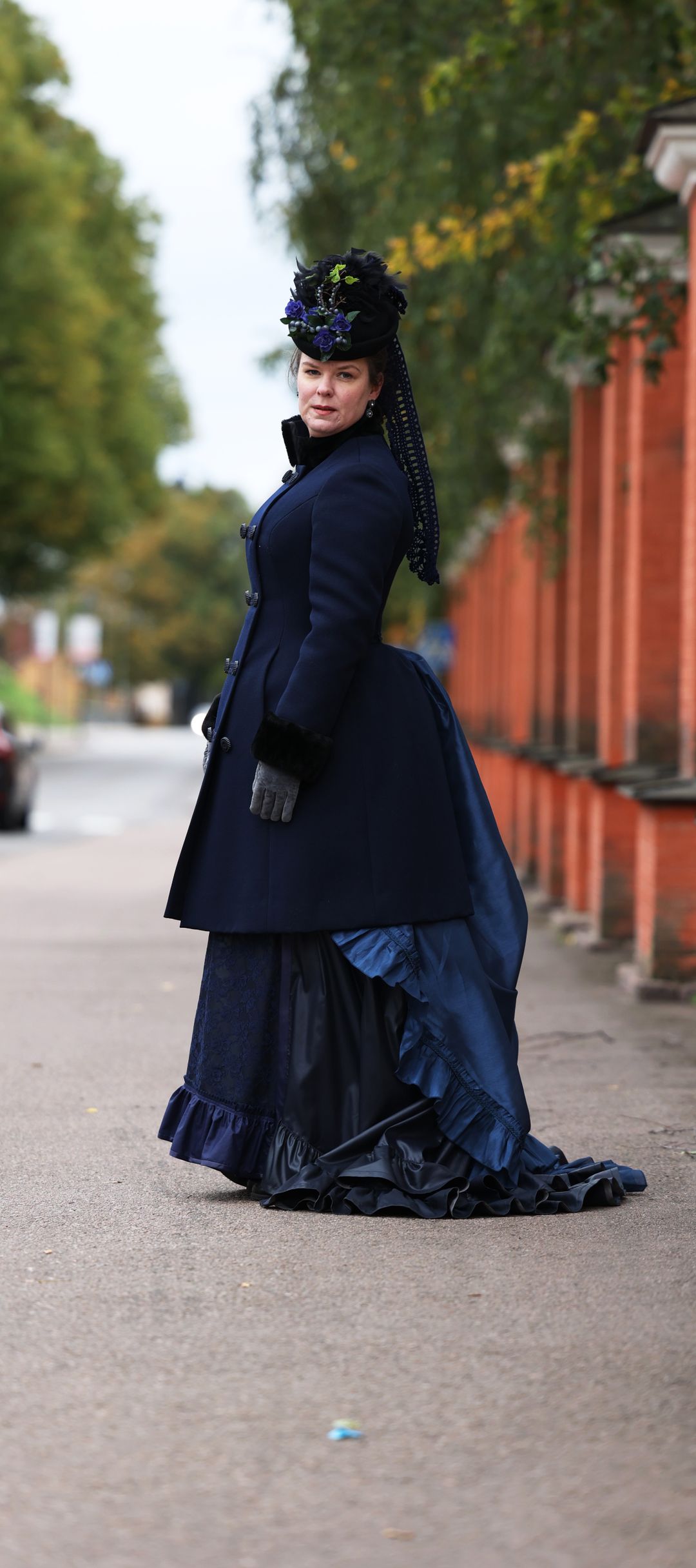 Taina Parikka seisoo 1880-luvun turnyyrilenignissä ja ulkotakissa kadulla Haminan ortodoksikirkon punertavan kiviaidan vieressä. Parikka seisoo sivuttain ja katsoo kameraan.