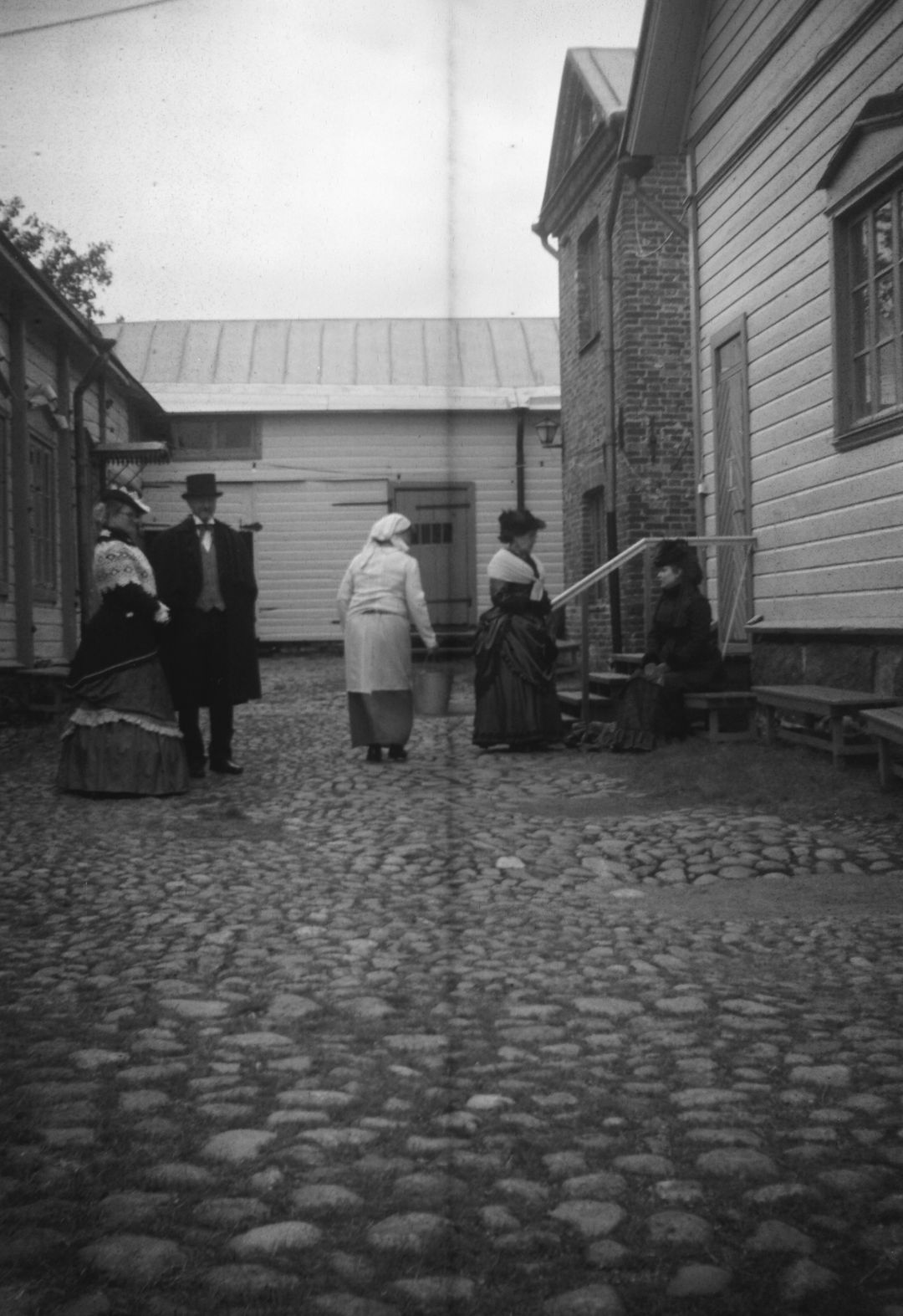 Mustavalkokuva Kauppiaantalomuseon sisäpihalta. Pihalla viisi Haminan Hywää Asukasta oleilee ja jutustelee 1800-luvun lopun asustuksissa.
