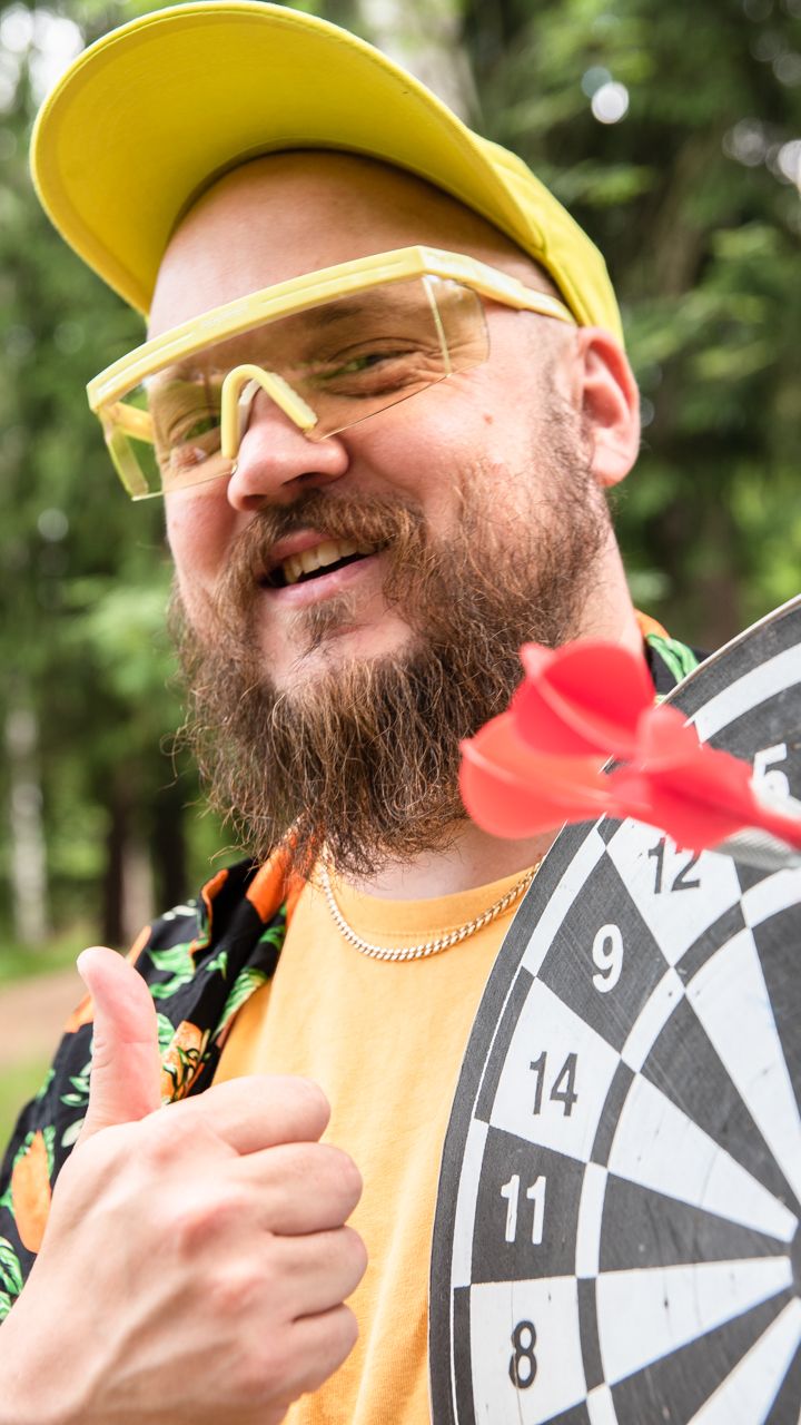 Rap-artisti Pyhimys katsoo hymyillen kameraan. Hän pitelee tikkataulua ja näyttää peukaloa. Taustalla on metsää.