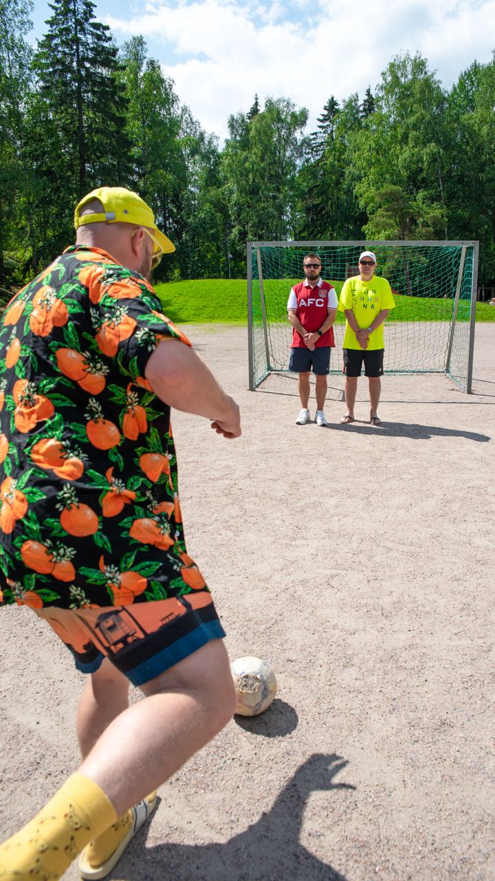 Pyhimys on selkä kohti kameraa, potkaisemassa palloa kohti edessään olevaa maalia. Maalin edessä seisovat rap-artistit Voli ja Heikki Kuula.