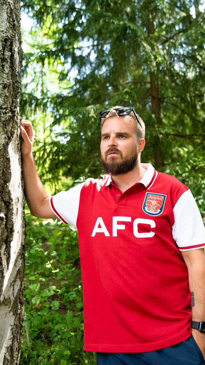 Voli nojaa puunrunkoon toisella kädellään ja katsoo jonnekin kauas. Hänellä on yllään punainen paita, ja otsalla aurinkolasit. Taustalla on metsää.
